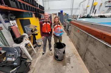 Two UNH student researchers hold their winning ocean renewable energy device in front of a wave tank. They are wearing red life jackets.